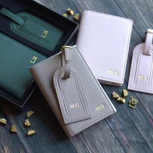 May include: A selection of personalised travel accessories, including passport holders and luggage tags in green, taupe, and pink. Each item features gold initials. The accessories are arranged on a dark wood surface.
