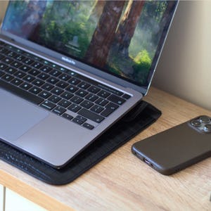 May include: A silver MacBook Pro laptop with a black keyboard sits on a black leather pad. A brown smartphone rests on a wooden surface next to the laptop. The laptop screen displays a nature scene.