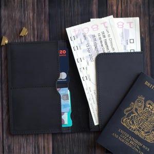 K&ouml;nnte beinhalten: Schwarzes Leder-Reise-Portemonnaie mit mehreren F&auml;chern, das einen Reisepass, Tickets und Kreditkarten enth&auml;lt. Das Portemonnaie ist ge&ouml;ffnet und zeigt seinen Inhalt auf einer dunklen Holzoberfl&auml;che.