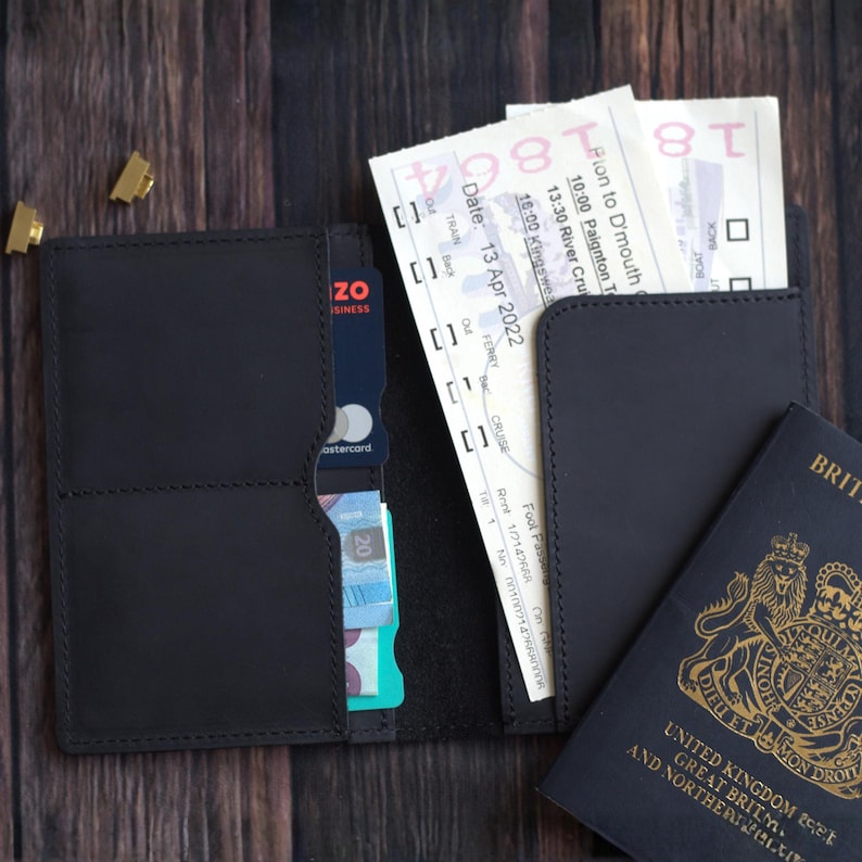 K&ouml;nnte beinhalten: Schwarzes Leder-Reise-Portemonnaie mit mehreren F&auml;chern, das einen Reisepass, Tickets und Kreditkarten enth&auml;lt. Das Portemonnaie ist ge&ouml;ffnet und zeigt seinen Inhalt auf einer dunklen Holzoberfl&auml;che.