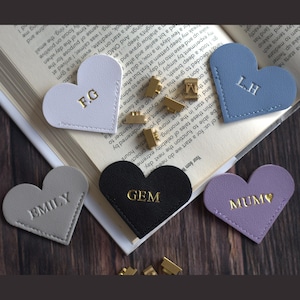 May include: Heart-shaped leather bookmarks in various colors, including gray, black, and lavender. Each bookmark features personalized gold lettering, such as initials or names. The bookmarks are arranged on an open book.