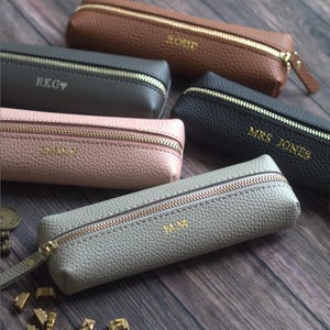 May include: A collection of personalized pencil cases in various colors, including brown, gray, pink, and black. Each case features a gold zipper and embossed initials or a name. The cases are made of textured leather-like material.