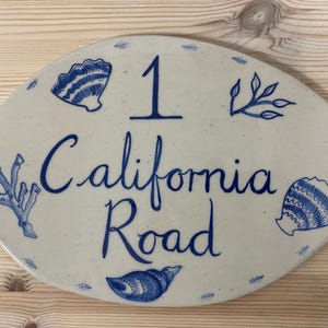 May include: Oval ceramic house sign featuring blue seashell and coral designs. The number "1" and the words "California Road" are written in blue script. The sign has two holes for mounting.