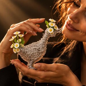 Puede incluir: Una escultura decorativa de alambre con forma de pato y acabado metálico plateado. La figura del pato presenta pequeñas flores de margarita blancas con centros amarillos en la cabeza y el lomo, integradas en la intrincada estructura de alambre.
