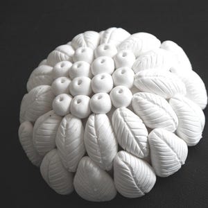Può includere: Una scultura bianca a forma di fiore con una superficie strutturata. La scultura è composta da forme arrotondate e forme a foglia, disposte in un motivo circolare. Lo sfondo è nero.