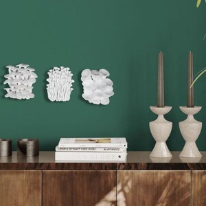 Puede incluir: Arreglo decorativo de arte mural con tres esculturas de hongos blancos. Dos candelabros de piedra con velas grises est&aacute;n sobre un mueble de madera. Dos peque&ntilde;os recipientes de metal y una pila de libros completan la decoraci&oacute;n contra una pared verde.