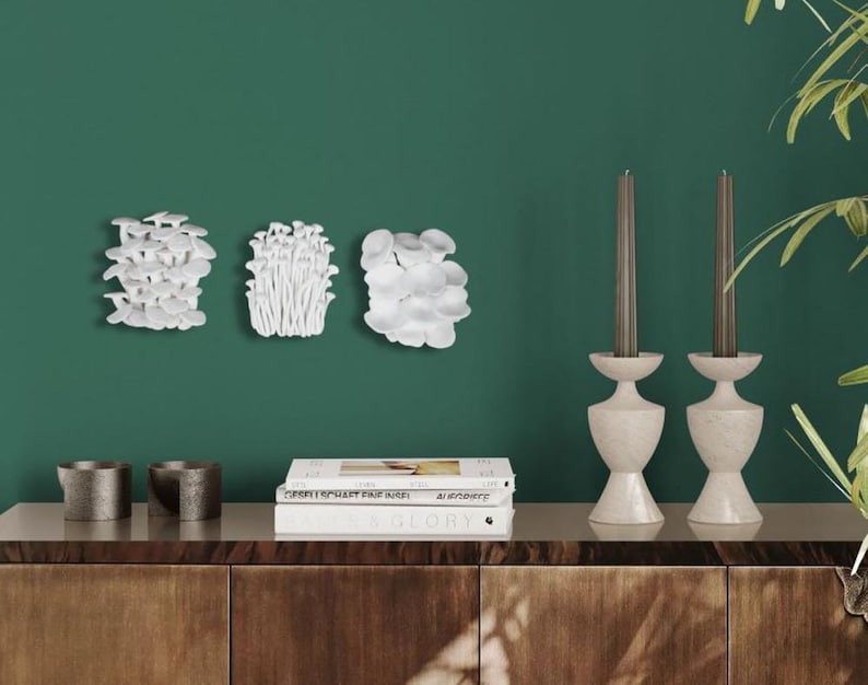 Puede incluir: Arreglo decorativo de arte mural con tres esculturas de hongos blancos. Dos candelabros de piedra con velas grises est&aacute;n sobre un mueble de madera. Dos peque&ntilde;os recipientes de metal y una pila de libros completan la decoraci&oacute;n contra una pared verde.