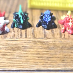 May include: A collection of small, colorful dragon figurines. The dragons are in various colors, including black with yellow accents, red and white, green and black, blue, and red and yellow. They are arranged on a wooden surface.