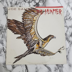 May include: Album cover for Joe Sample's "The Hunter." The cover features a detailed illustration of a hawk in flight, with brown and tan feathers and a sharp beak. The title is in red, stylized lettering.