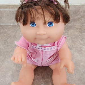 Può includere: Una bambola vintage con capelli castani acconciati in due codini, ciascuno fissato con un fiocco rosa. La bambola ha gli occhi blu e indossa una tutina rosa a quadretti vichy con finiture floreali bianche. La bambola è seduta.