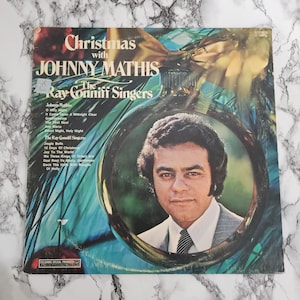 May include: A vintage vinyl record album cover featuring Johnny Mathis, with the text "Christmas with Johnny Mathis" and "The Ray Conniff Singers." The album art includes a portrait of Johnny Mathis inside a Christmas ornament.