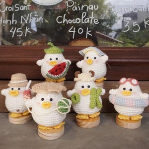 Puede incluir: Seis figuras de patos de ganchillo hechas a mano, cada una con accesorios únicos como sombreros y bufandas. Los patos son blancos con picos y patas amarillas, presentados sobre bases de madera. El fondo incluye texto escrito a mano.