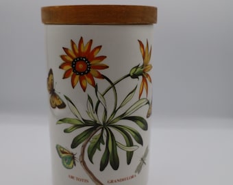 May include: A white ceramic canister with a wooden lid, featuring botanical illustrations of orange and yellow flowers, green leaves, and butterflies. The canister has the text "ARCTOTIS GRANDIFLORA".