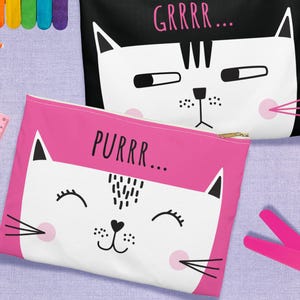 May include: Two zippered pencil cases with cat faces. One is black with "GRRRR..." in pink, the other is pink with "PURRR..." in black. The cat faces are white with black details and pink cheeks. A pink ruler, scissors, and glue are also visible.