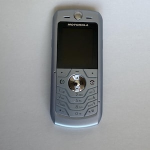 Op de afbeelding: Een zilverkleurige Motorola mobiele telefoon met een rechthoekig zwart scherm, een centraal rond navigatiepaneel met een metallic afwerking en een standaard numeriek toetsenbord met in het zwart gedrukte alfanumerieke tekens.