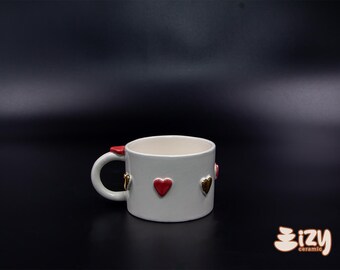 May include: White ceramic mug with a handle and red and gold heart embellishments. The mug has a smooth, glossy finish and a simple, elegant design. The mug is perfect for enjoying your favorite hot beverages.