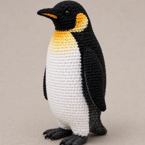 Puede incluir: Un pingüino de ganchillo hecho a mano en negro, blanco, amarillo y gris. El pingüino tiene una cabeza negra con detalles amarillos, una barriga blanca y pies grises. El pingüino está de pie.