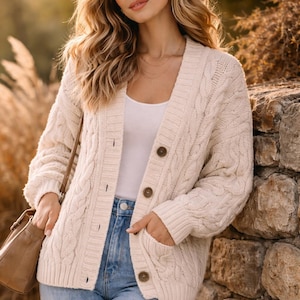 May include: Cream cable knit cardigan with brown buttons and pockets, worn with a white vest and light blue jeans. A brown handbag is visible. The cardigan is a classic knitwear piece.