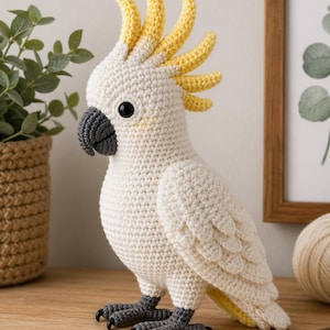 May include: A handmade crochet cockatoo bird in cream and yellow. The bird has a distinctive yellow crest, black eyes, and a gray beak and feet. The bird is standing on a wooden surface.