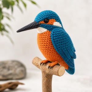 May include: A handmade crochet kingfisher bird with a blue head and wings, an orange breast, and a long black beak. The bird is perched on a small wooden branch, showcasing its detailed craftsmanship. The bird's eyes are black, and it has white accents.