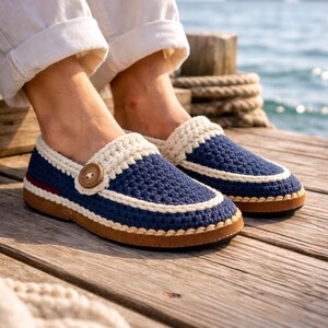 May include: Navy blue crochet slippers with a cream-colored trim and a wooden button detail. The slippers have a brown sole and a red stripe accent. These handmade slippers are perfect for indoor wear.