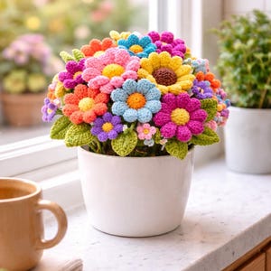 Peut inclure: Un bouquet vibrant de fleurs crochetées dans un pot en céramique blanc. Les fleurs sont de différentes couleurs, dont le rose, le bleu, l'orange et le jaune, avec des feuilles vertes. Le pot est sur un comptoir blanc près d'une fenêtre.