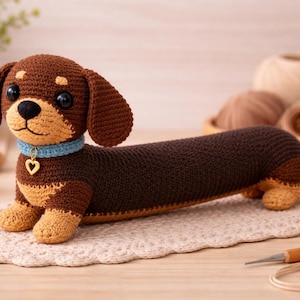 Op de afbeelding: Een gehaakte teckel in bruin en beige met een blauwe halsband en hartvormige bedel. De handgemaakte hond ligt op een kanten kleedje, met garen en een haaknaald op de achtergrond. Een charmant, handgemaakt item.
