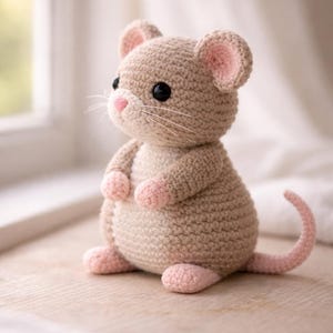 May include: A handmade crochet mouse toy in beige with pink accents. The mouse has large black eyes, a pink nose, and a long pink tail. The ears and paws are also pink. The toy is sitting on a wooden surface.