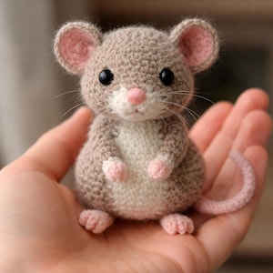 May include: A handmade crochet mouse toy in shades of tan, white, and pink. The mouse has large black eyes, a pink nose, and a long, curled tail. The toy is held in a person's hand.