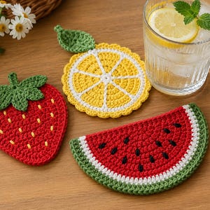 May include: Crocheted fruit-shaped coasters. A red strawberry, a yellow lemon slice, and a watermelon slice with black seeds and a green rind are displayed. The coasters are handmade and sit on a wooden surface.