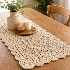 Könnte beinhalten: Beige gehäkelter Tischläufer mit gewelltem Rand auf einem Holztisch. Eine Vase mit Grün, Kerzen und ein Holzuntersetzer sind auf dem Läufer platziert. Das offene Webmuster des Läufers ist sichtbar.