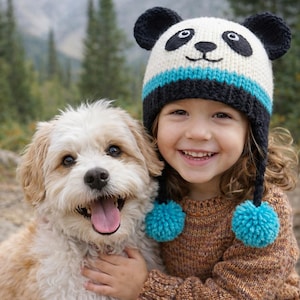 May include: A smiling child wearing a panda bear knit hat with blue pom-poms, hugging a fluffy, light-colored dog. The child is wearing a brown sweater. The background is a blurred outdoor scene.