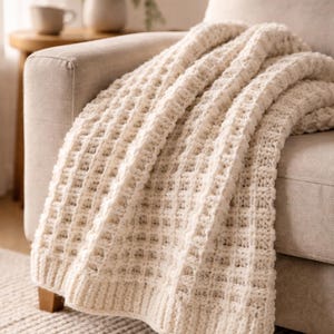 Könnte beinhalten: Cremefarbene Strickdecke, die über einem beigen Sofa drapiert ist. Die Decke hat ein strukturiertes, waffelartiges Muster. Ein kleiner Beistelltisch aus Holz mit einer Tasse und einer Topfpflanze befindet sich im Hintergrund.