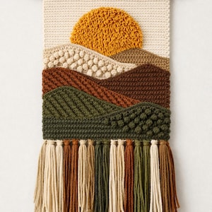 Puede incluir: Un tapiz de pared artesanal con un diseño de paisaje texturizado. Presenta un sol amarillo, colinas en capas en tonos marrones y verdes, y una franja de borlas. La obra de arte está montada en una varilla de madera.