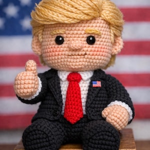 May include: Crocheted doll of a man in a black suit, red tie, and white shirt, with a small American flag pin. The doll has blonde hair and is giving a thumbs-up. The background features an American flag.