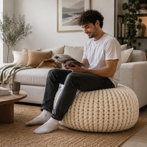 May include: A cream-colored, knitted pouf in a living room setting. A person is sitting on the pouf, reading a magazine. The room features a neutral color palette with a beige sofa, a wooden coffee table, and a jute rug.