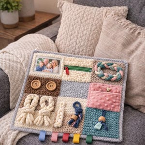 Peut inclure: Tableau sensoriel fait main avec diverses textures et activités. Comprend un cadre photo, une fermeture éclair, des boutons, une corde tressée, des rubans et des poches. Le tableau est principalement crème, bleu et marron, conçu pour l'exploration tactile et l'apprentissage précoce.