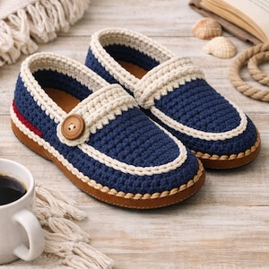 Puede incluir: Un par de zapatillas azules hechas a mano con detalles en crema y un botón de madera. Las zapatillas tienen una suela marrón y una raya roja en el lateral. Se muestran sobre una superficie de madera.