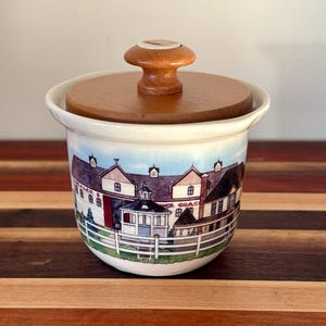 May include: A white ceramic jar with a round wooden lid featuring a knob handle. The jar is decorated with a colorful illustration of a multi-building farm complex, a white picket fence, and the text "DEALER & COACH" printed on the structure.