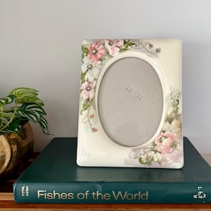 May include: A rectangular ceramic picture frame with an oval center opening, featuring a cream-colored base decorated with hand-painted pink and white floral motifs, green leaves, and delicate vine accents along the top and bottom corners.