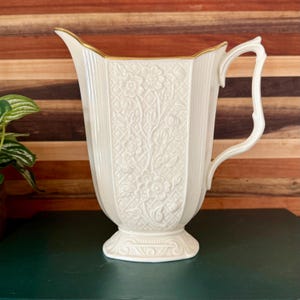 May include: A cream-colored ceramic pitcher featuring an embossed floral pattern over a lattice background. The vessel has a gold-trimmed rim, a fluted body, a pedestal base with decorative molding, and a curved handle on the right side.