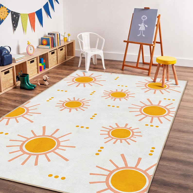 May include: A white rug with a repeating pattern of orange and peach suns. The rug is in a child's playroom, with a wooden easel, a small yellow stool, and a white chair. A shelf with books and toys is visible.