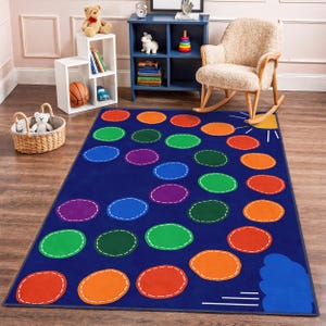 Op de afbeelding: Een blauw rechthoekig tapijt met een kleurrijk hinkelbaanontwerp. Het heeft ovale vormen in rood, oranje, groen, paars en blauw, omlijnd met witte stiksels. Een gele zon en een blauwe wolk staan in de hoek. Geschikt voor een kinderkamer.