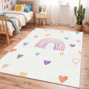Könnte beinhalten: Ein rechteckiger, weißer Teppich mit einem Aquarell-Regenbogen und bunten Herzen. Der Regenbogen hat rosa, lila und blaue Bögen. Herzen in Orange-, Gelb-, Lila- und Blautönen sind auf dem Teppich verstreut. Ein rosa Rand umrahmt den Teppich.