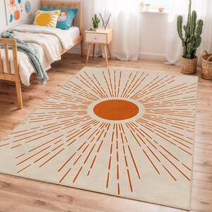 Könnte beinhalten: Ein rechteckiger Teppich mit cremefarbenem Hintergrund und einem großen orangefarbenen Sonnenmuster. Der Sonnenstrahl geht von einem zentralen orangefarbenen Oval aus, mit Linien, die sich nach außen erstrecken. Der Teppich liegt auf einem Holzboden.