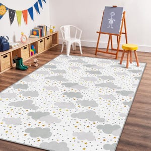 Puede incluir: Alfombra rectangular con fondo blanco, con un diseño de nubes grises, estrellas doradas y puntos. La alfombra está colocada sobre un suelo de madera en una sala de juegos infantil, con una silla blanca, un caballete y estanterías.