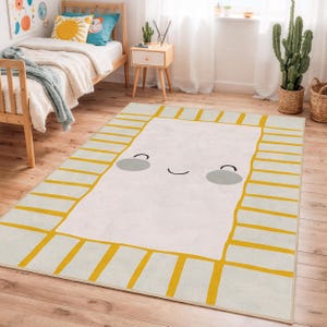 Puede incluir: Una alfombra rectangular con un diseño de cara sonriente. La alfombra tiene un centro color crema con óvalos grises para los ojos y una línea curva para la boca. El borde es crema con rayas amarillas.