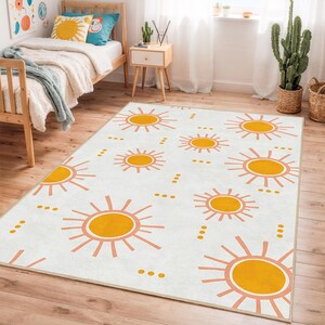 May include: A rectangular area rug with a repeating pattern of stylized yellow suns with orange rays on a white background. The rug is placed on a wooden floor in a room with a bed and a side table.