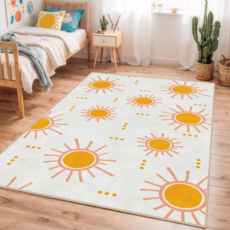 May include: A rectangular area rug with a repeating pattern of stylized yellow suns with orange rays on a white background. The rug is placed on a wooden floor in a room with a bed and a side table.