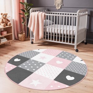 Puede incluir: Una alfombra redonda para guardería con un diseño de patchwork. La alfombra presenta cuadrados rosas, grises y blancos, con lunares, corazones y estrellas. Una cuna blanca con una manta rosa está en el fondo.